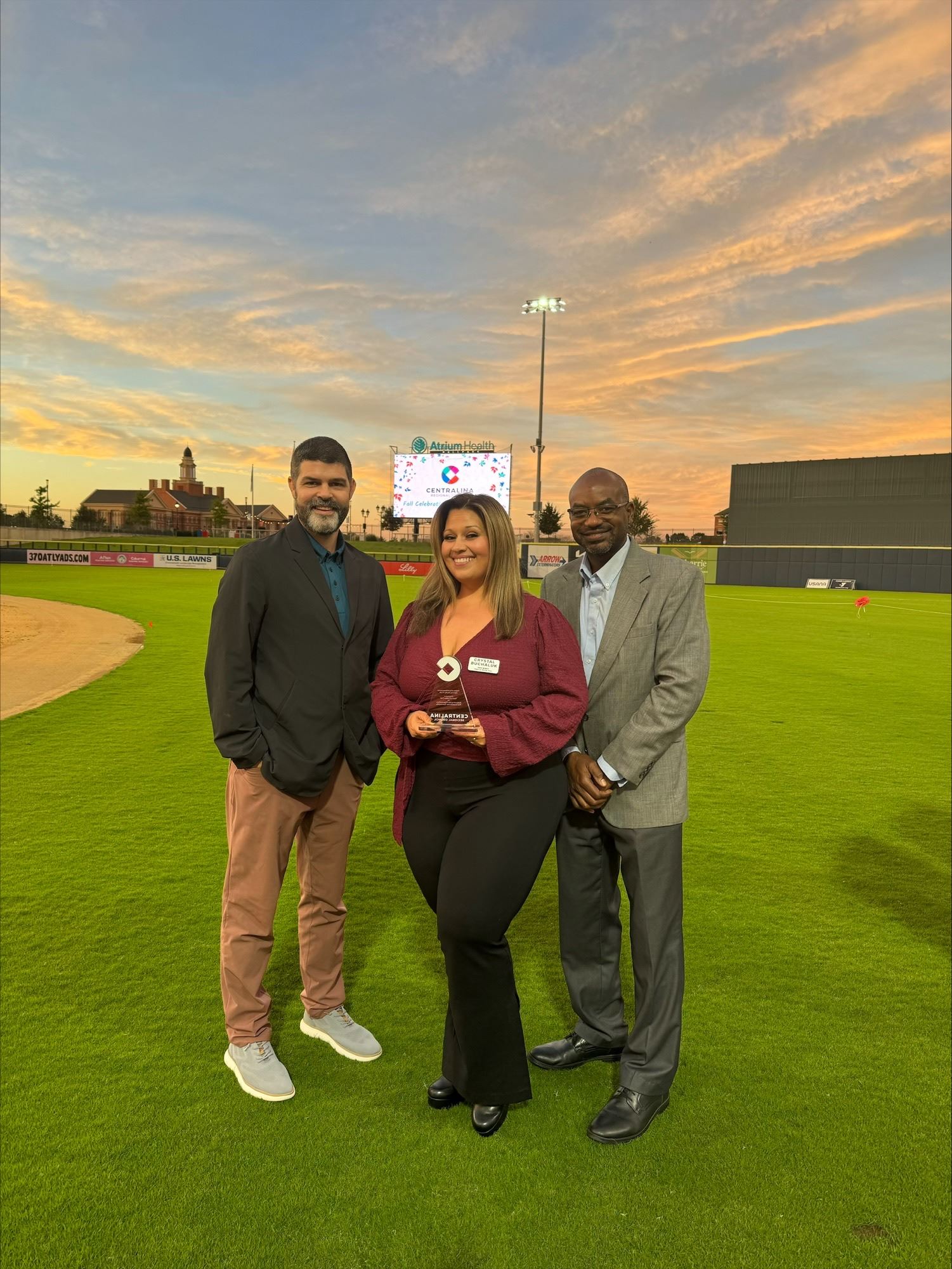 Three people stand on a baseball field with a sunset behind them. One person is holding an award.