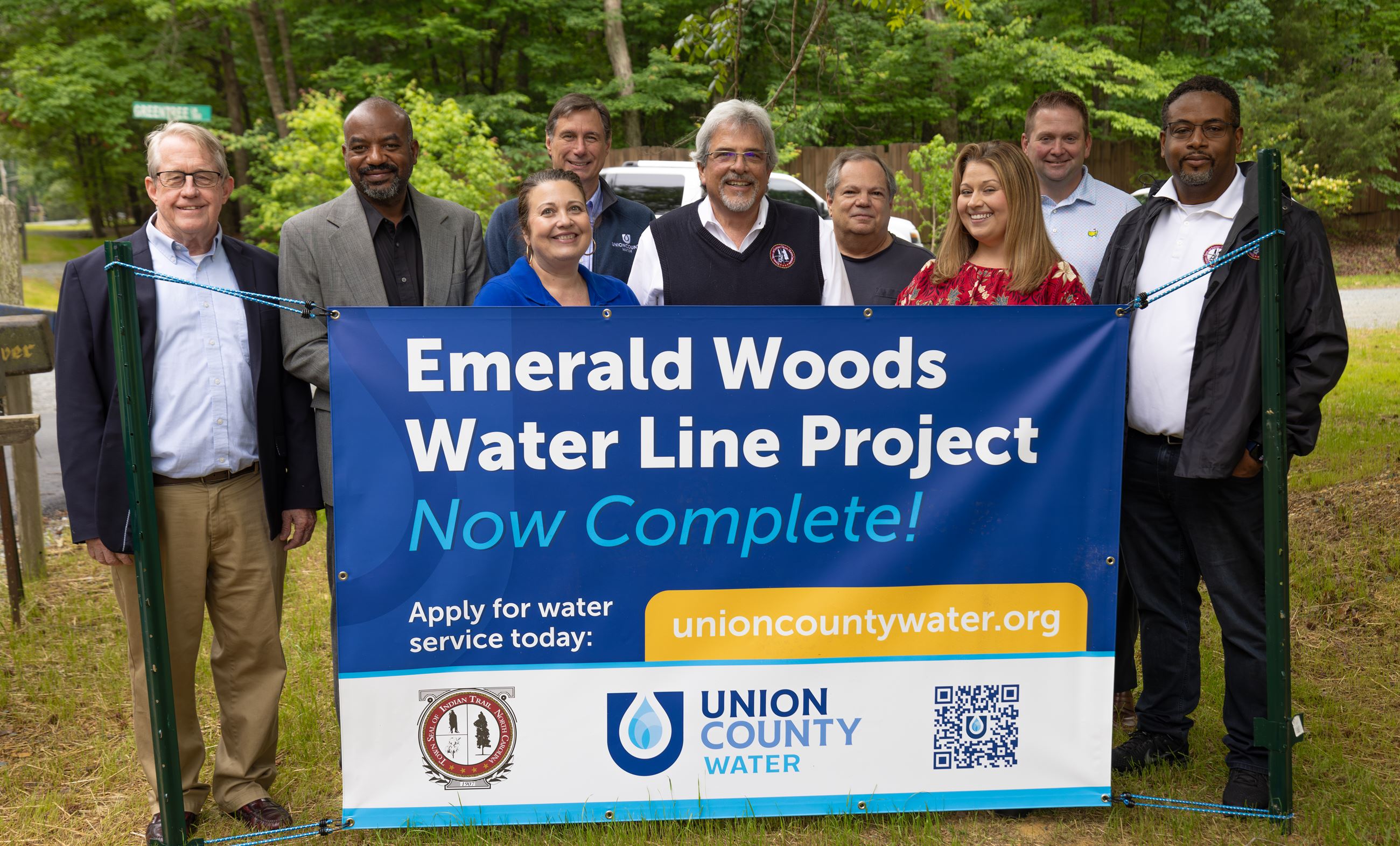 A group of 9 men and women stand behind a banner celebrating a waterline project. 